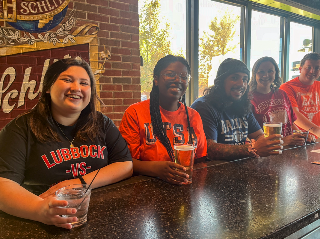 Five smiling people sit at a bar with drinks, wearing college t-shirts that say Lubbock, UTSA, Dallas, Oklahoma, and FAU. Sunlight streams through large windows behind them.